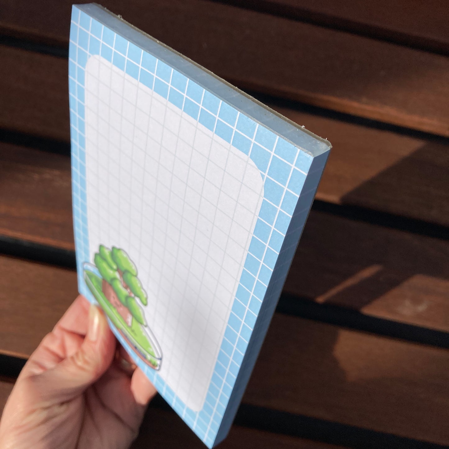 A hand holding the notepad at an angle to show the short top glued edge and a side edge of the notepad.