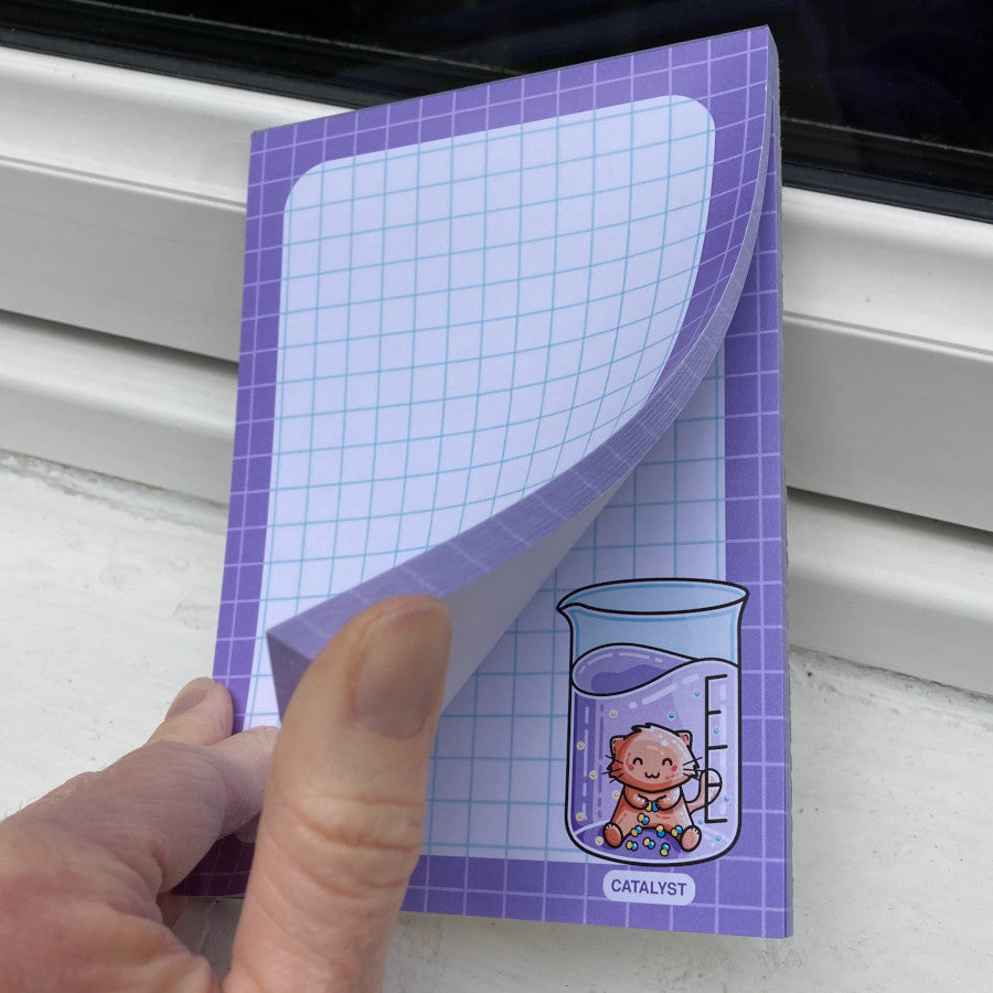 A hand flicking through the sheets of a white notepad with a purple border and blue graph lines featuring a fun catalyst pun design in the corner.