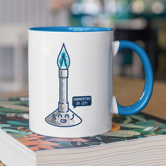 A blue and white two toned ceramic mug standing on a book with the handle to the right, and a design of a cute Bunsen burner with blue flame and speech bubble saying chemistry is lit!