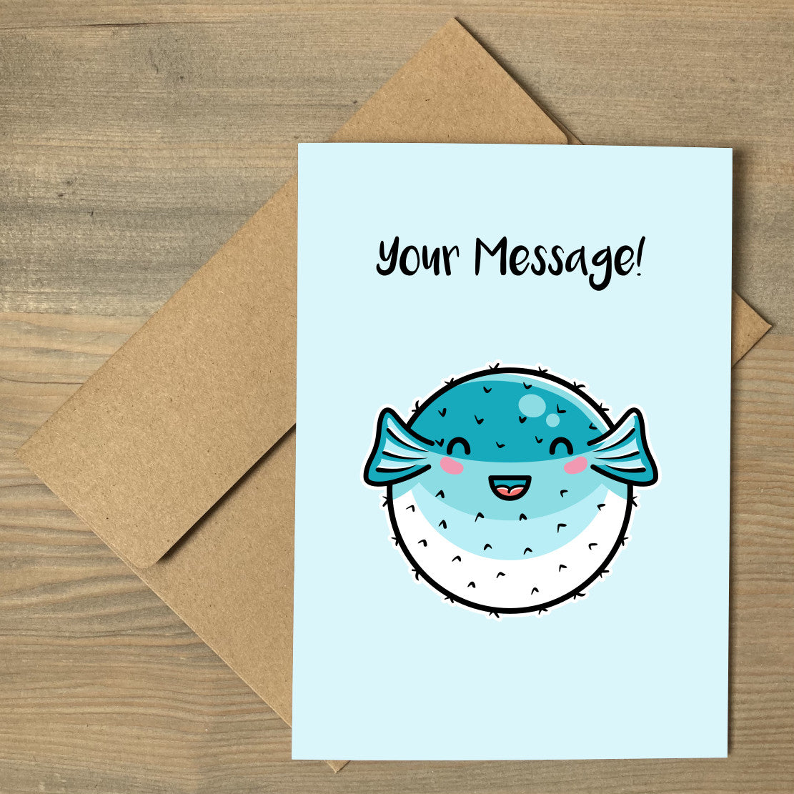 A pale turquoise greeting card lying flat on a brown envelope, with a design of a kawaii cute turquoise and white puffer fish with personalised wording above