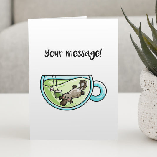 A white greeting card standing on a white table, with a design of a cute platypus swimming in a glass teacup of green tea with personalised wording above