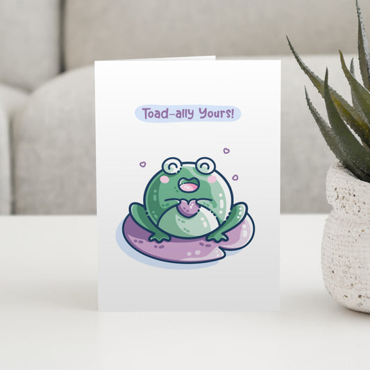 A white greeting card standing on a table and featuring a kawaii cute green toad holding a purple heart and sitting on a purple lilly pad. The words toad-ally yours appear above the toad.
