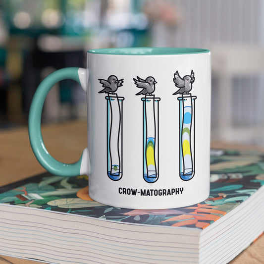 A two toned white and spearmint ceramic mug with the handle to the left, standing on a thick book, showing a design of 3 crows holding strips of paper into 3 test tubes showing colour separation.