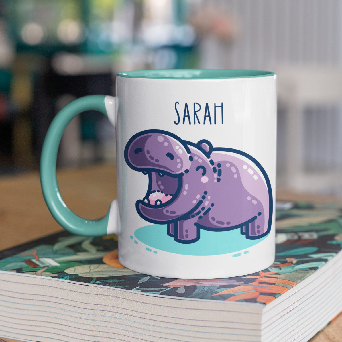 A white ceramic mug with a spearmint handle on the left and yellow inside. A name is written in thin dark blue upper case letters above a kawaii cute purple hippo with a thick dark blue outline and a turquoise shadow beneath. The hippo is seen side on facing to the left and looks happy with its mouth open wide.