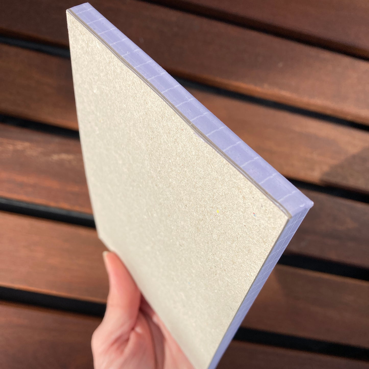 A hand holding the notepad at an angle to show the brown greyboard back of the notepad.