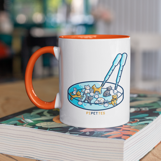 A white mug with an orange handle and inside, standing on a book. Design on the mug is a petri dish or cats and rabbits with two pipettes and the word pipettes beneath.