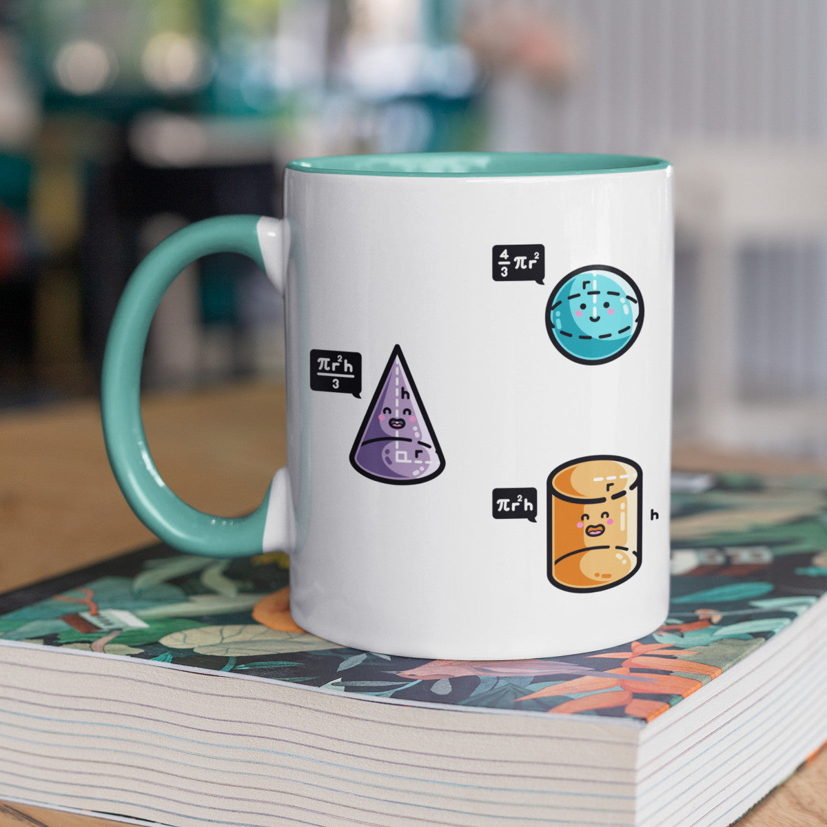 A two toned white and spearmint ceramic mug with the handle to the left showing three colourful 3D shapes with faces and speech bubbles stating the equation for working out their volume