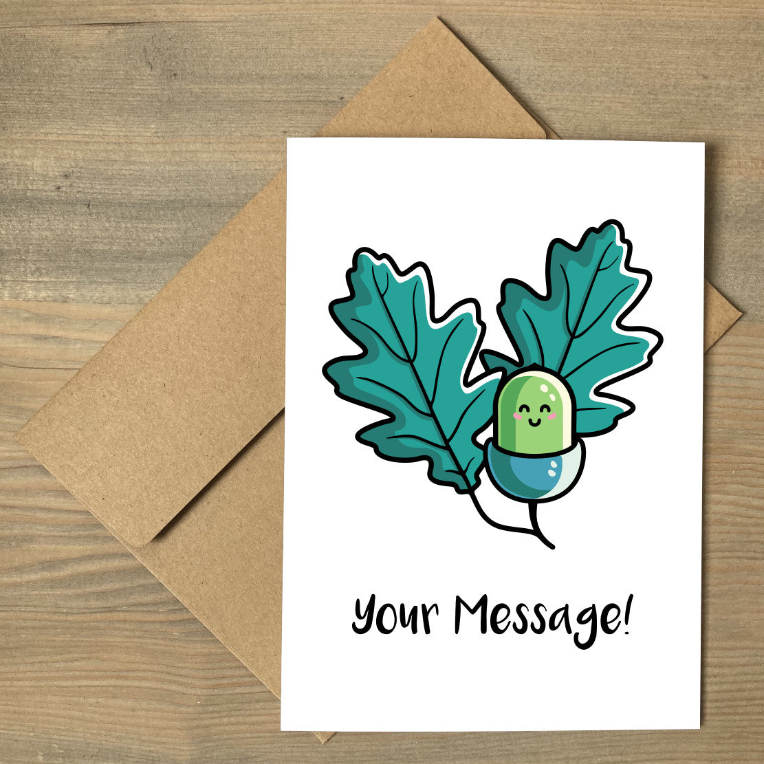 A brown envelope beneath a white greeting card with a design of a kawaii cute green acorn with two oak leaves behind on either side and the words your message written beneath