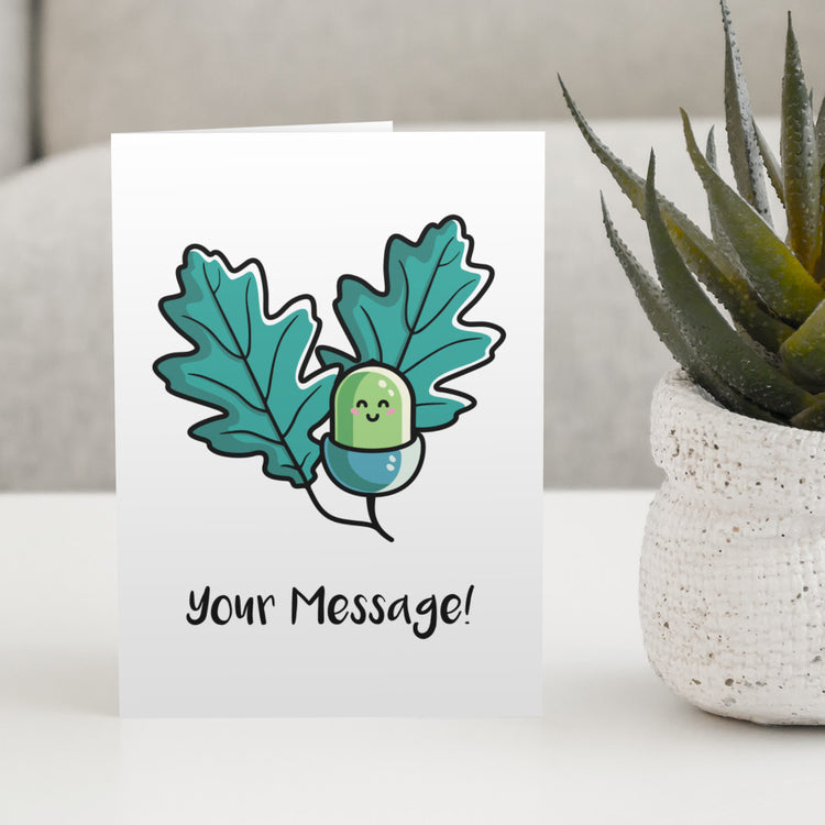A white greeting card standing on a white table with a design of a kawaii cute green acorn with two oak leaves behind on either side and the words your message written beneath
