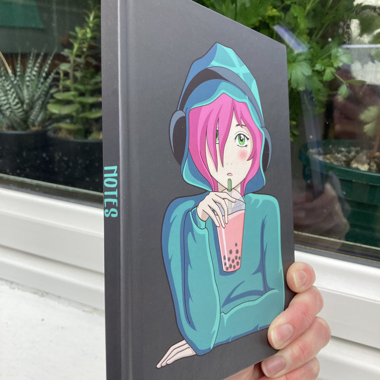 Dark grey hardback journal held in a hand side on showing the word notes written on the spine and an anime girl with pink hair on the front