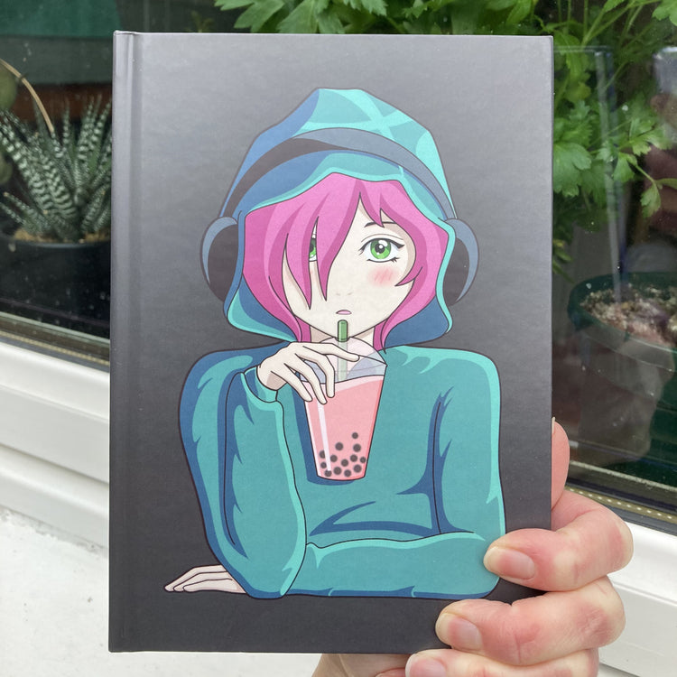 Grey hardback journal held in a hand showing the front with a picture of an anime girl with pink hair and green eyes wearing a green hoodie and headphones drinking boba
