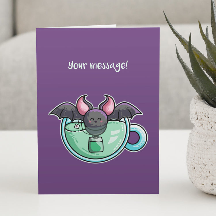 A purple greeting card standing on a white table with a design of a cute bat in a cup of green tea with a personalised message above.