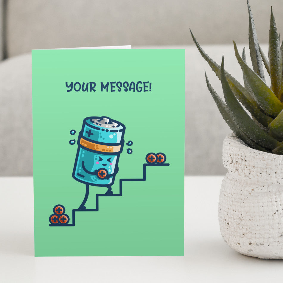 A green greeting card standing on a white table next to a plant, the card has a design of a kawaii cute cylindrical blue battery working hard, sweating, carrying a positive charge up some steps and the words your message written above