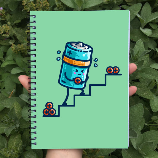 Held in a hand is a closed spiral notebook showing a pale green front cover with a design of a kawaii cute cylindrical blue battery working hard, sweating, carrying a positive charge up some steps.