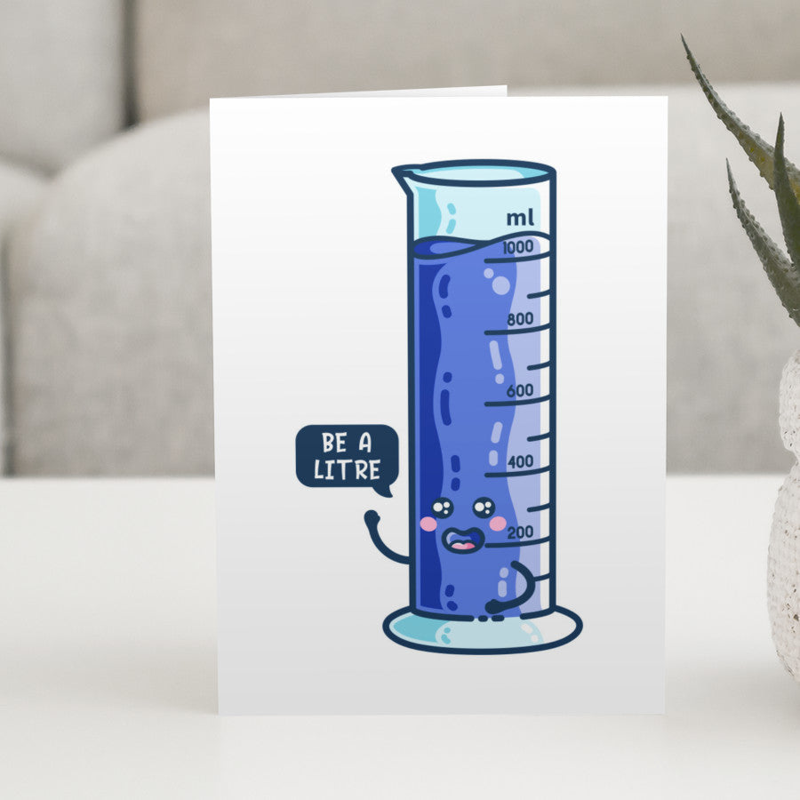 A white greeting card standing on a white table, with a design of a happy blue one litre graduated cylinder with a speech bubble saying be a litre