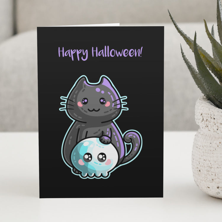 A black greeting card standing on a white table with a design of a kawaii cute black cat with its paw on a blue skull with a personalised message above.