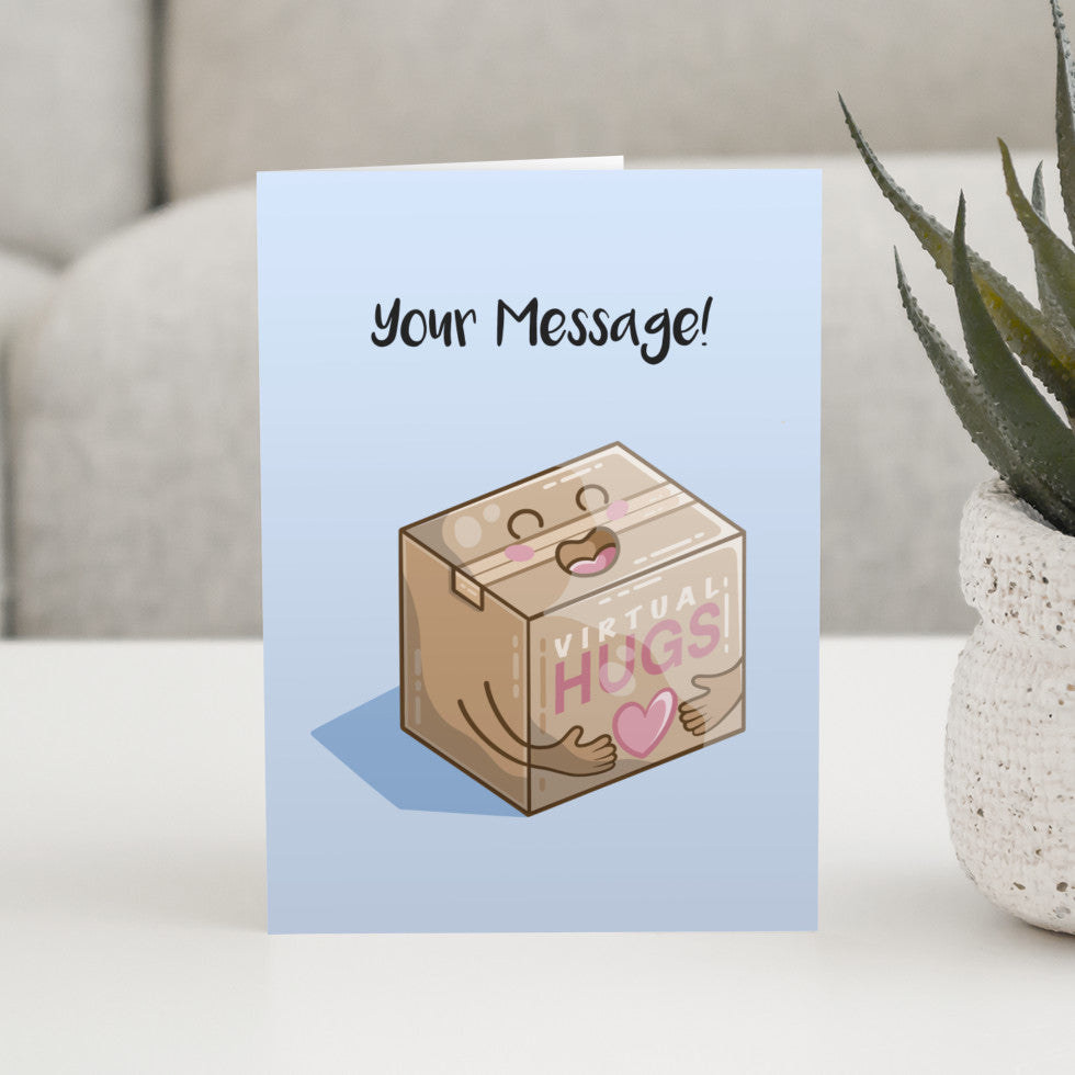 A pale blue greeting card standing on a white table with a design of a kawaii cute brown cardboard box with the words VIRTUAL HUGS on it and a big smile, heart and hugging arms and the words your message written above