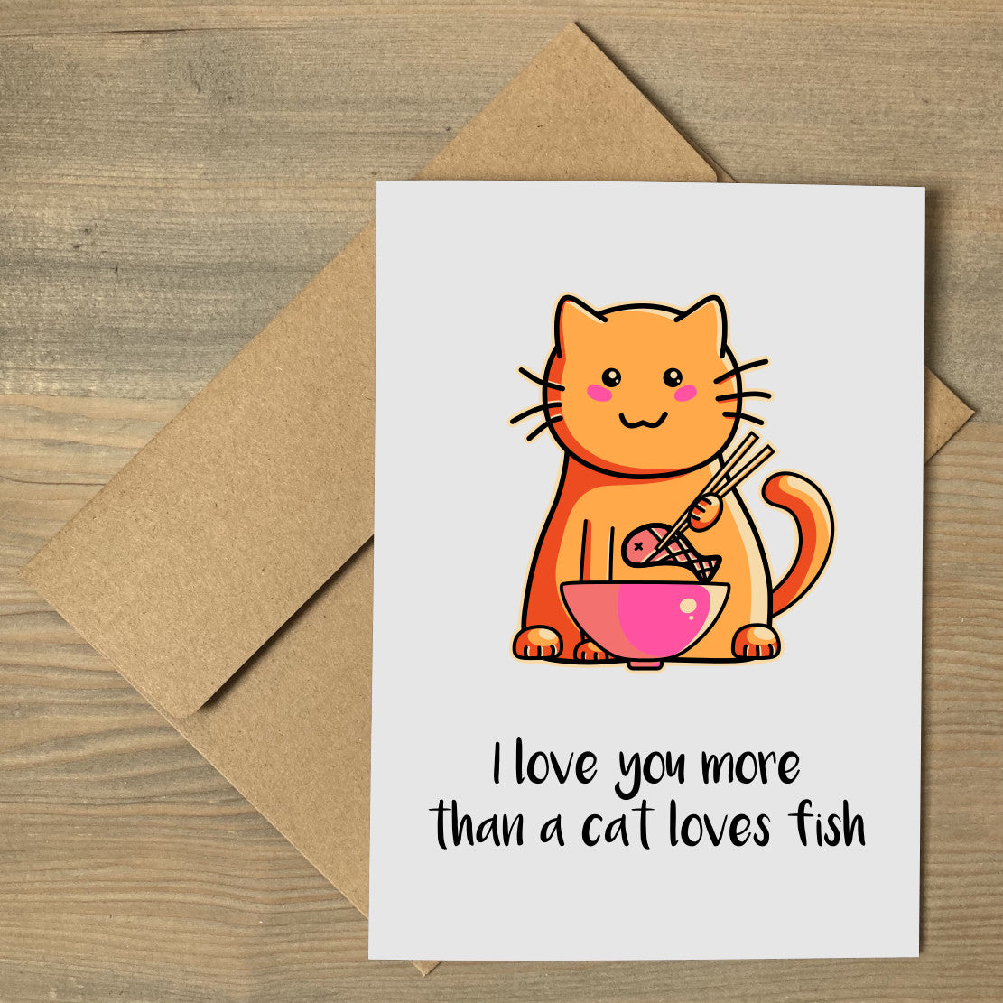 A grey greeting card lying flat on a brown envelope, with a design of a cute ginger cat eating a fish out of a bowl with chopsticks and with the words I love you more than a cat loves fish beneath