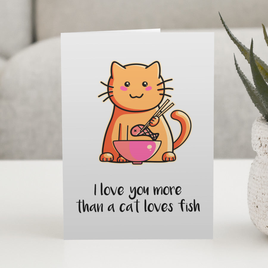A grey greeting card standing on a white table, with a design of a cute ginger cat eating a fish out of a bowl with chopsticks and with the words I love you more than a cat loves fish beneath