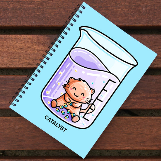 Blue notebook with black spiral binding lying on wooden slats, notebook design is of a cute ginger cat in a chemistry beaker and the word catalyst