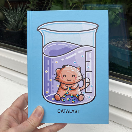 Blue hardback journal held showing the front with a design of a chemistry beaker containing purple liquid and a cat acting as a catalyst joining yellow and blue atoms together and the word catalyst written in capital letters beneath