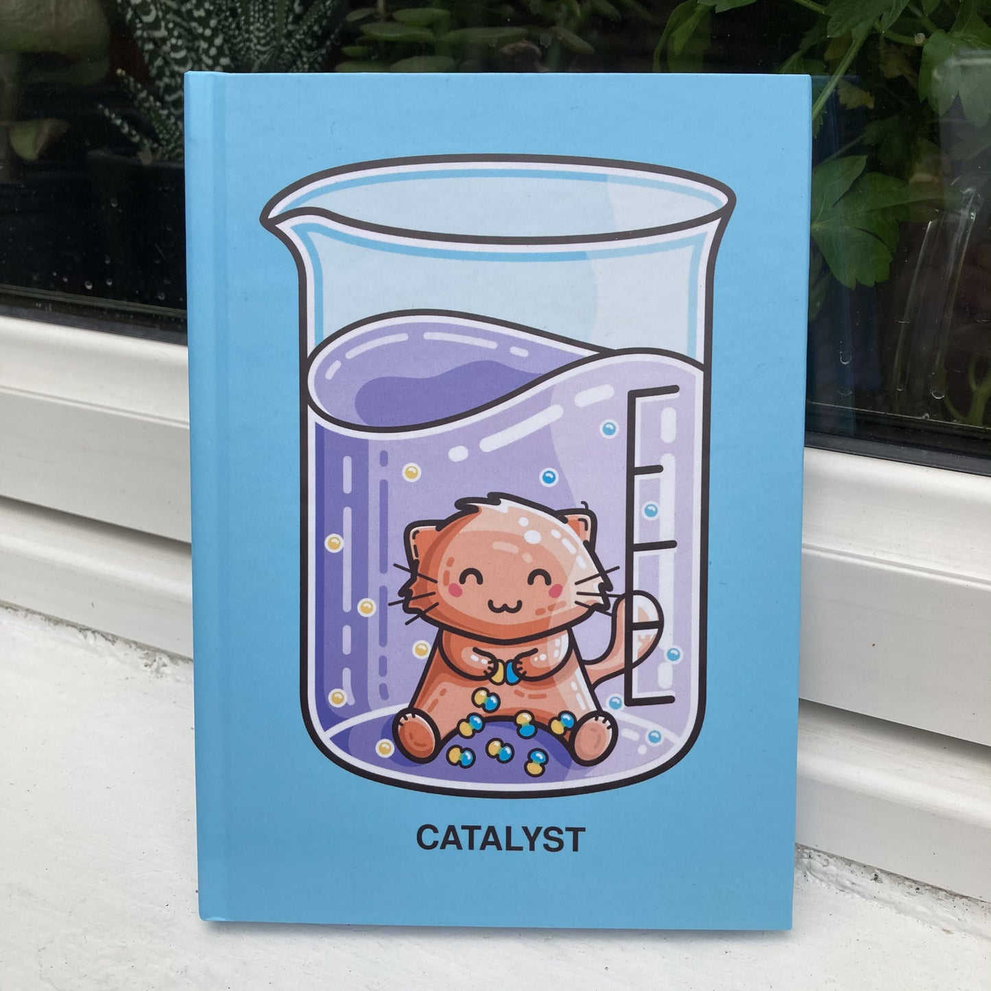 Blue hardback journal standing showing the front with a design of a chemistry beaker containing purple liquid and a cat acting as a catalyst joining yellow and blue atoms together and the word catalyst written in capital letters beneath