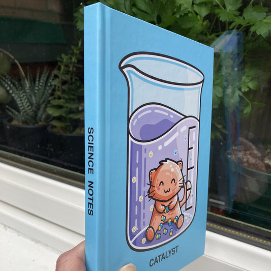 Blue hardback journal held at an angle showing the words science notes written on the spine and a cute cat in a beaker catalyst pun design on the front