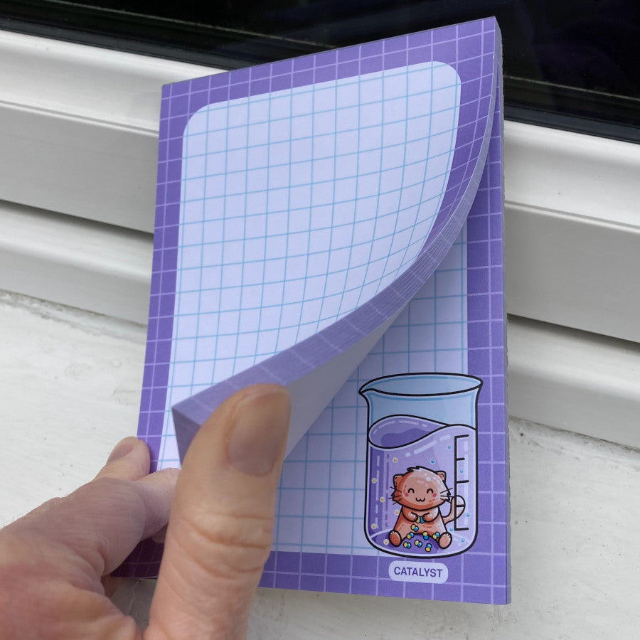 A hand flicking through the sheets of a white notepad with a purple border and blue graph lines featuring a fun catalyst pun design in the corner.