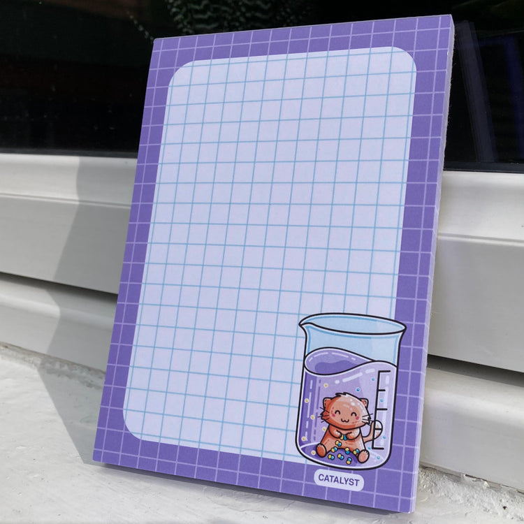White notepad with a purple border and blue graph lines. In the bottom right hand corner is a design of a cat acting as a catalyst in a chemistry beaker.