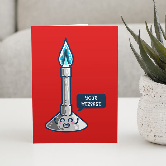 A red greeting card standing on a white table next to a plan. The card has a design of a kawaii cute bunsen burner lit with a blue flame and a speech bubble with the words your message to the right