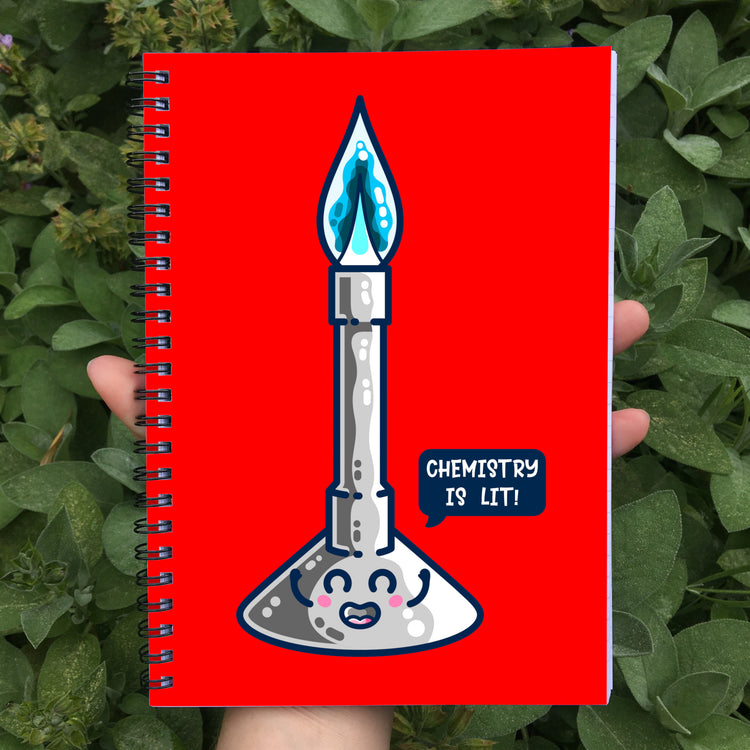 Held in a hand is a closed spiral notebook showing red front cover with a design of a cute Bunsen burner with a blue flame at the top and a smiling face on the base with a speech bubble saying chemistry is lit! The spiral wire is black.