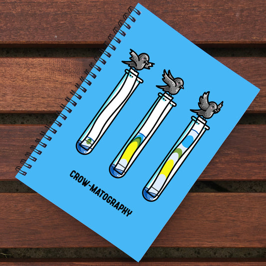 Blue notebook with black spiral binding lying on wooden slats, notebook design is of 3 cute crows holding strips of paper into 3 test tubes showing colour separation.