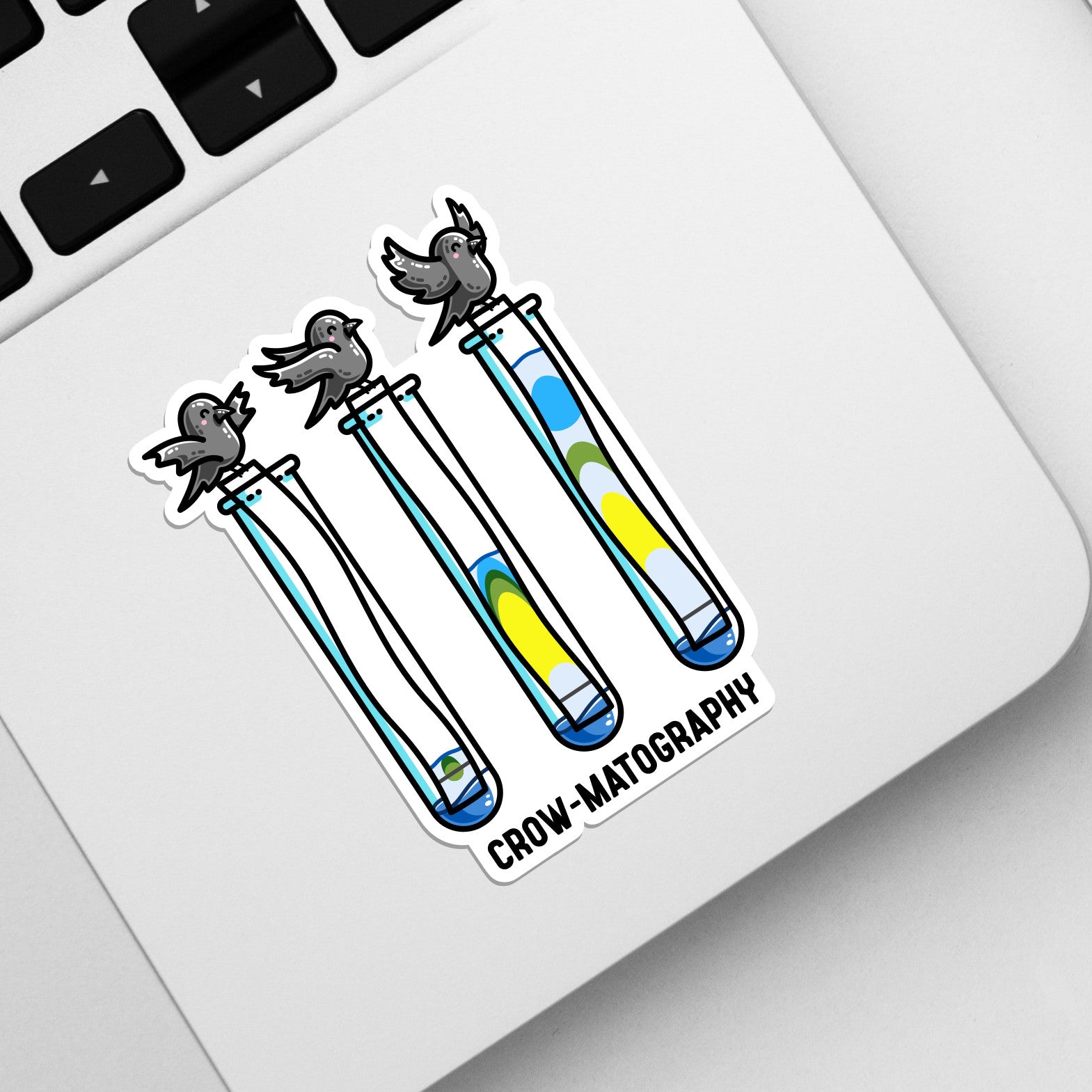 The corner of a laptop keyboard with a die cut vinyl sticker on it. The sticker is of 3 crows holding strips of paper into 3 test tubes showing colour separation, and the pun word crow-matography..