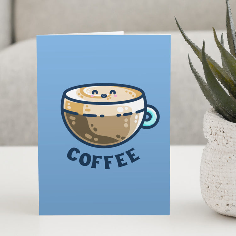A blue greeting card standing on a white table next to a plant and with a design on the card of a kawaii cute glass cup of coffee with a thick creamy layer at the top and a smiling face in the cream on the top. The word coffee is in capital letters beneath.