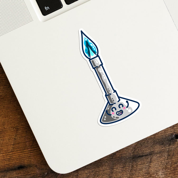 Photo of a vinyl die cut sticker in the shape of a bunsen burner stuck onto the left hand corner of a laptop computer keyboard. The sticker is a digital drawing of a kawaii cute bunsen burner with a blue flame and a happy smiling face on its base.