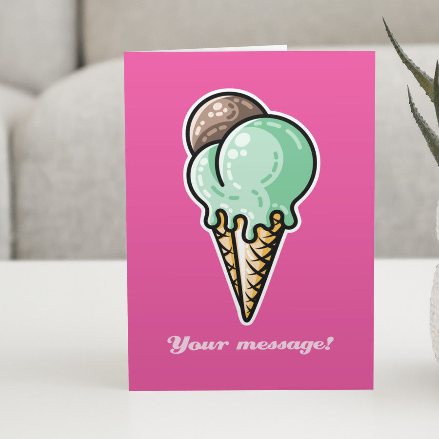 A pink greeting card standing on a white table, with a design of a cone with three scoops of ice cream, two green and one brown, and a personalised message below
