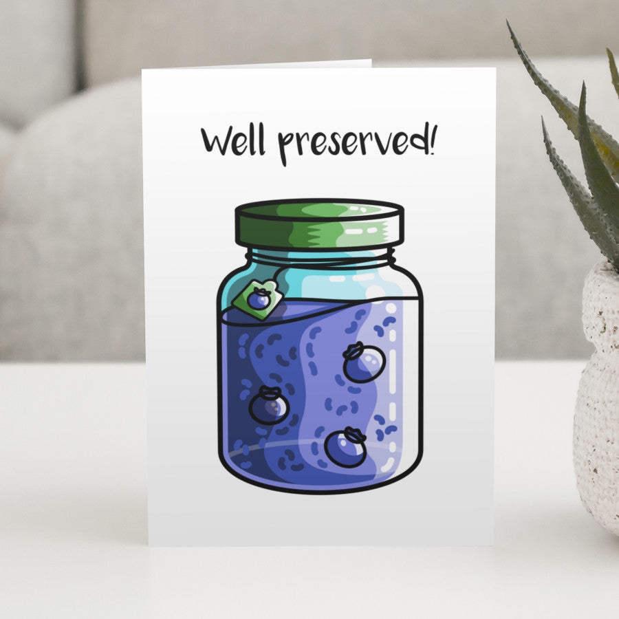 A white greeting card standing on a white table with a design of a jar of blueberry jam with a green lid with a personalised message above.