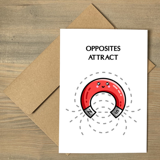 A white greeting card lying flat on a brown envelope, with the wording 'opposites attract' above a design of a cute red horseshoe shaped magnet and field lines