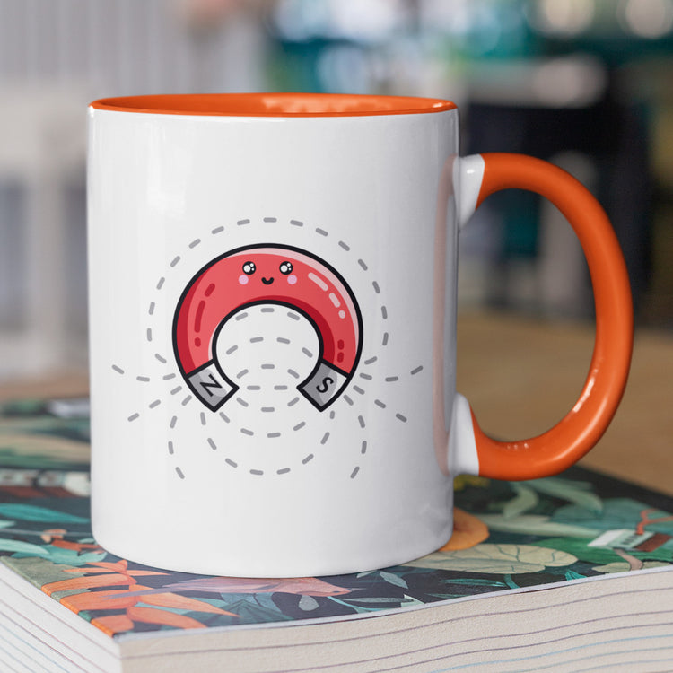 A white and orange two toned ceramic mug standing on a book, with the handle to the left, and a design of a cute red magnet with magnetic field lines.