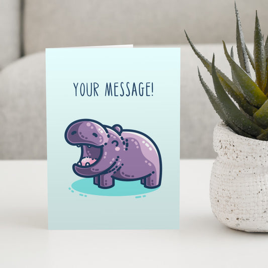 A large pale turquoise greeting card standing on a white table next to a plant. On the card is a kawaii cute purple hippo with a thick dark blue outline, seen side on facing to the left with its happy mouth open wide. There is a dark turquoise shadow beneath the hippo and thin upper case dark blue text above saying 'your message!'