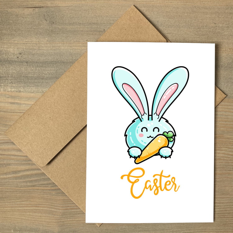 A white greeting card lying flat on a brown envelope, featuring a design of a kawaii cute pale pastel turquoise coloured rabbit holding a carrot like a pen with the word Easter written beneath in orange