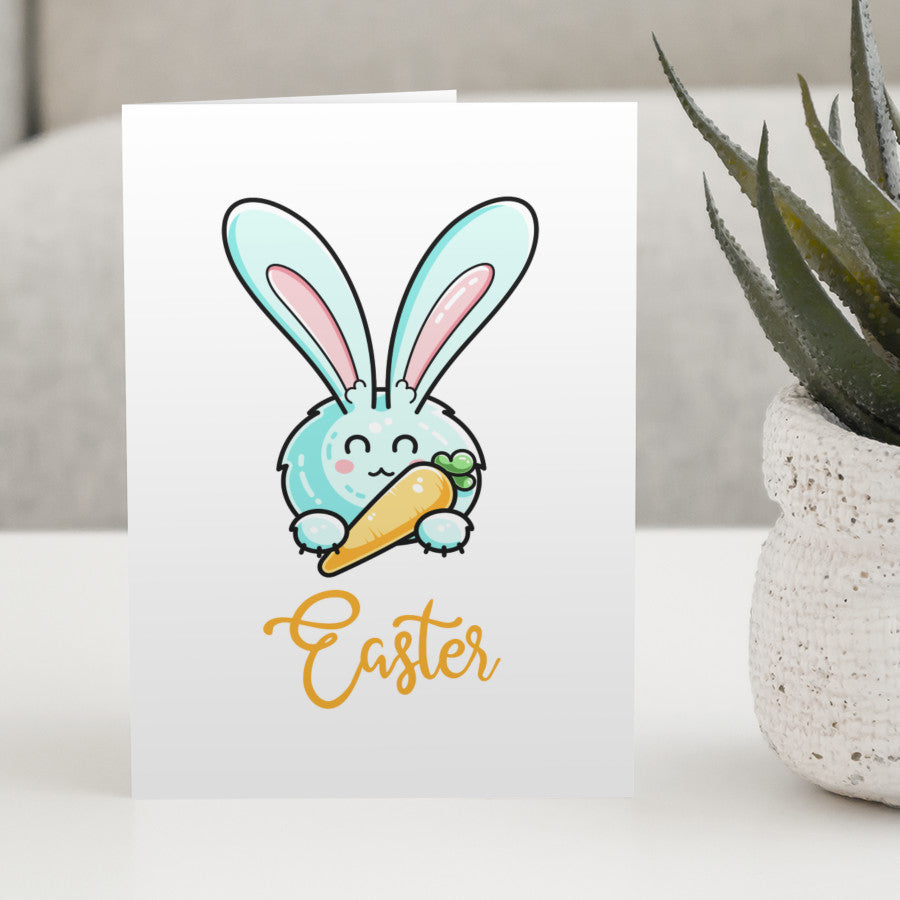 A white greeting card standing on a white table, featuring a design of a kawaii cute pale pastel turquoise coloured rabbit holding a carrot like a pen with the word Easter written beneath in orange
