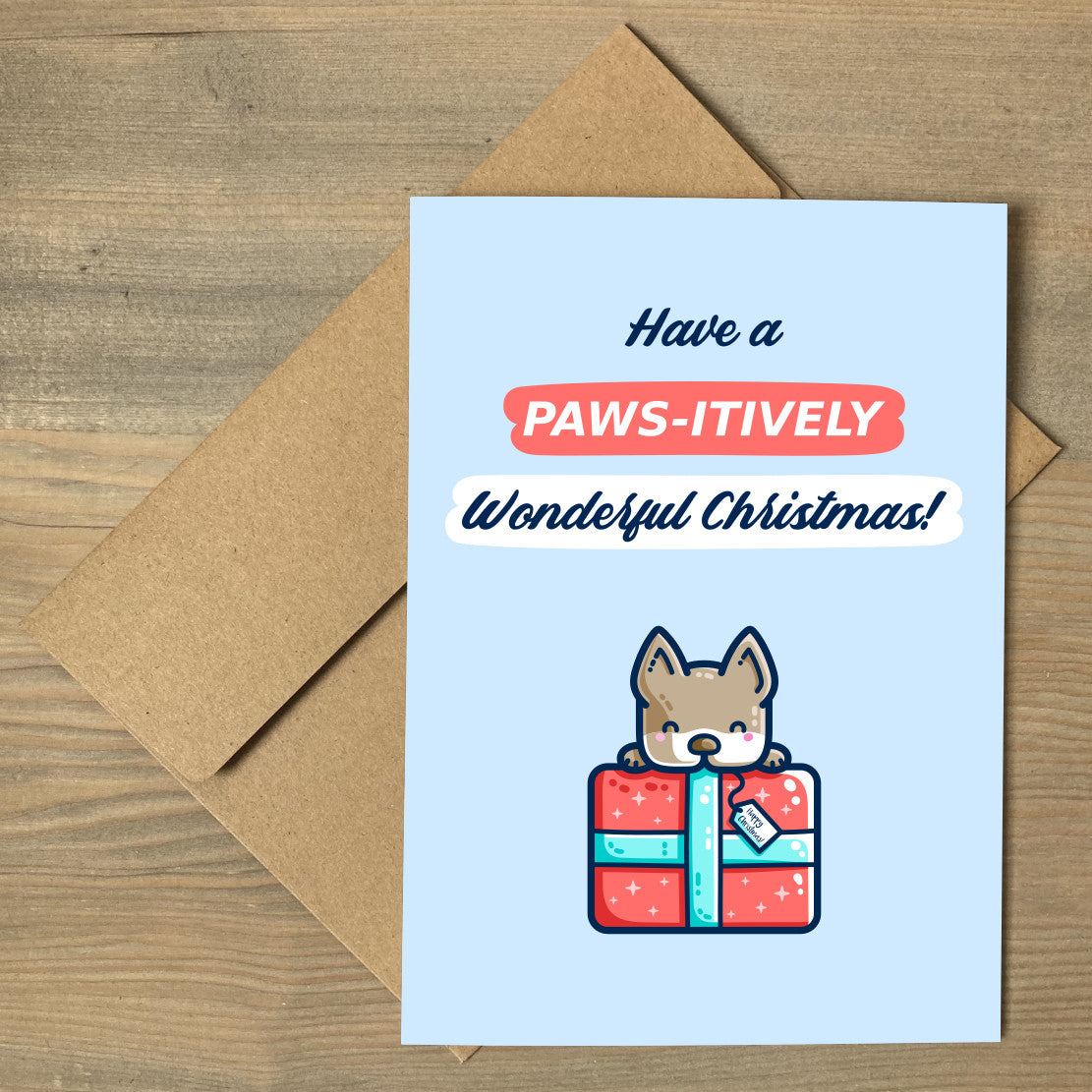 A pale blue card with the words 'have a paws-itively wonderful Christmas!' written above a picture of a dog peering over the top of a large wrapped red present. The card is lying flat on a brown envelope.