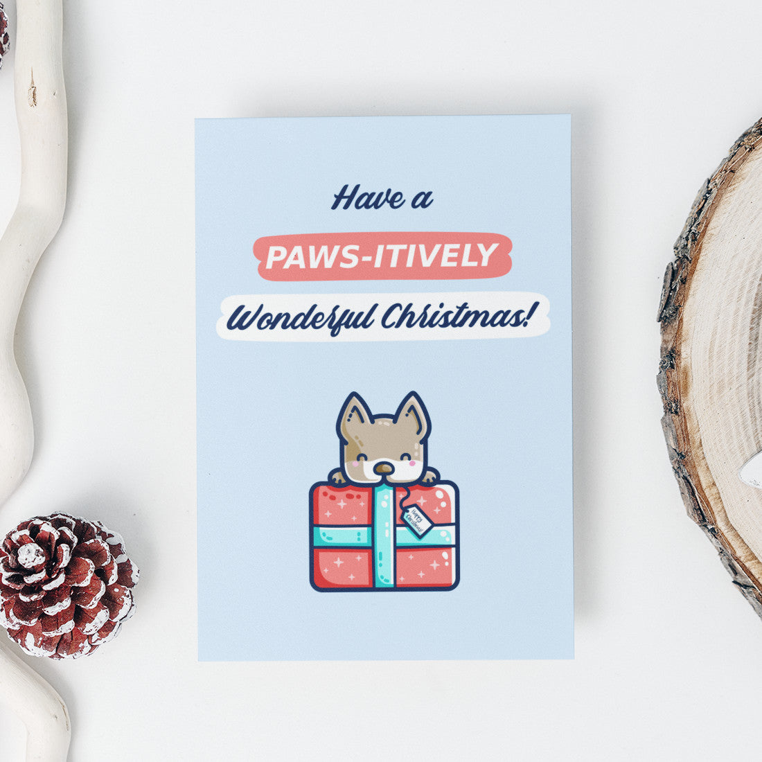 A pale blue card with the words 'have a paws-itively wonderful Christmas!' written above a picture of a dog peering over the top of a large wrapped red present. The card is lying flat on a white background with festive decorations around it.