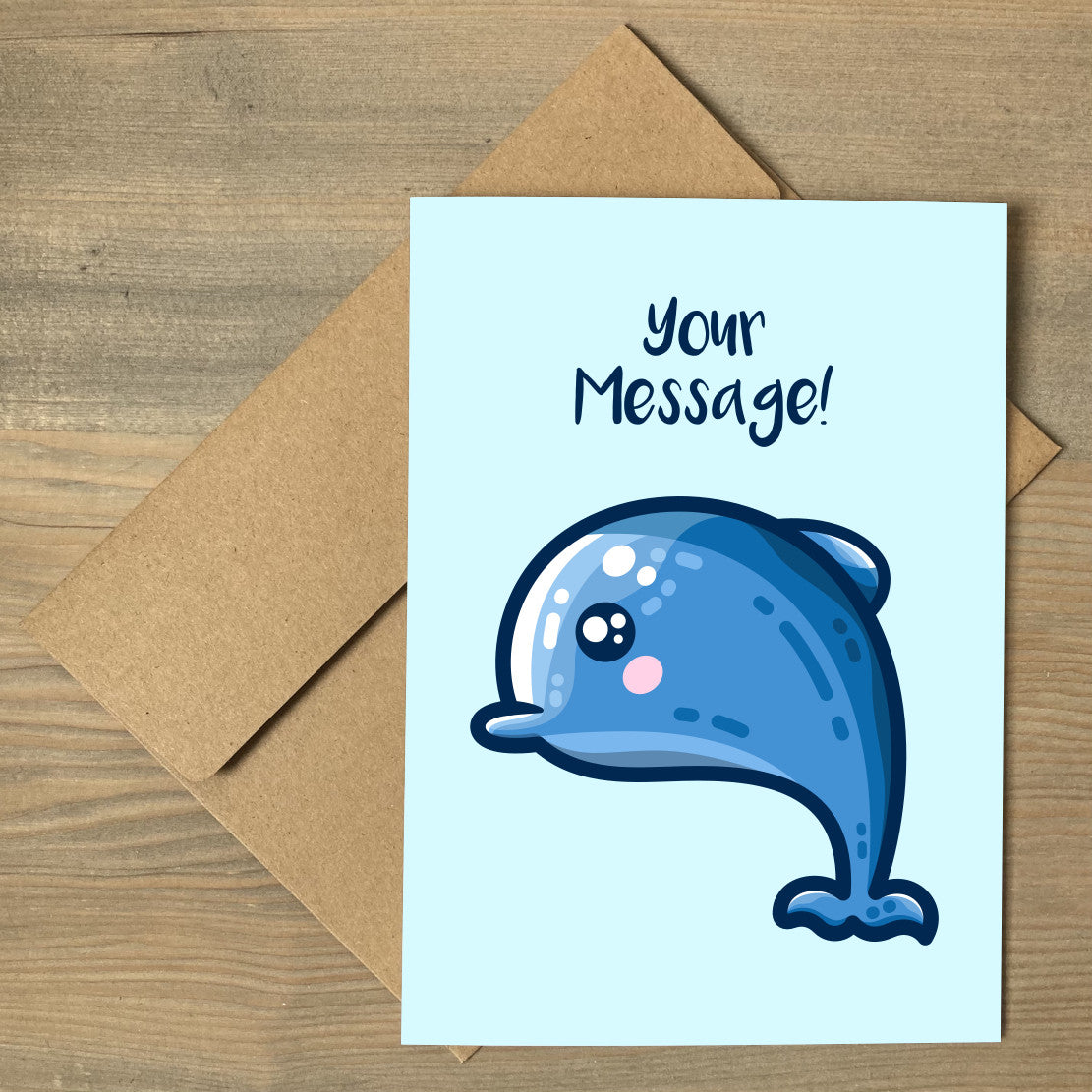 A pale blue greeting card lying flat on a brown envelope with a design of a kawaii cute blue dolphin and the personalised words Your Message written above