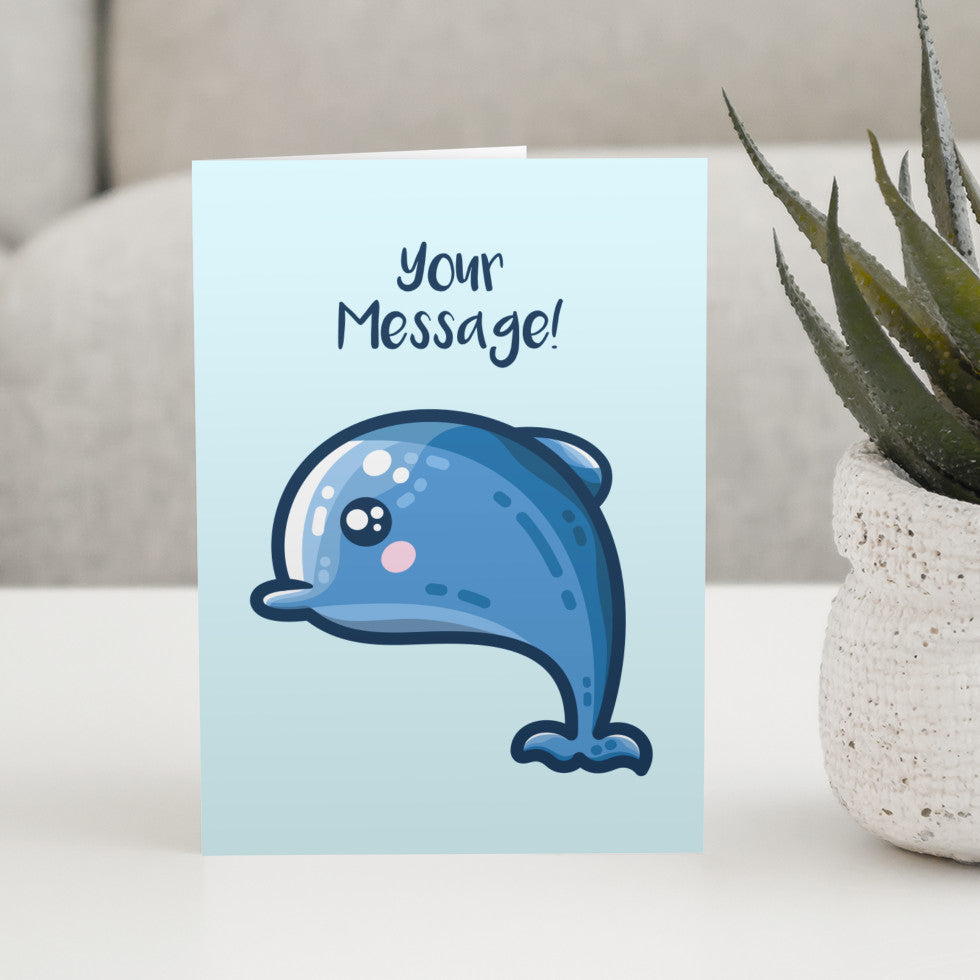 A pale blue greeting card standing on a white table with a design of a kawaii cute blue dolphin