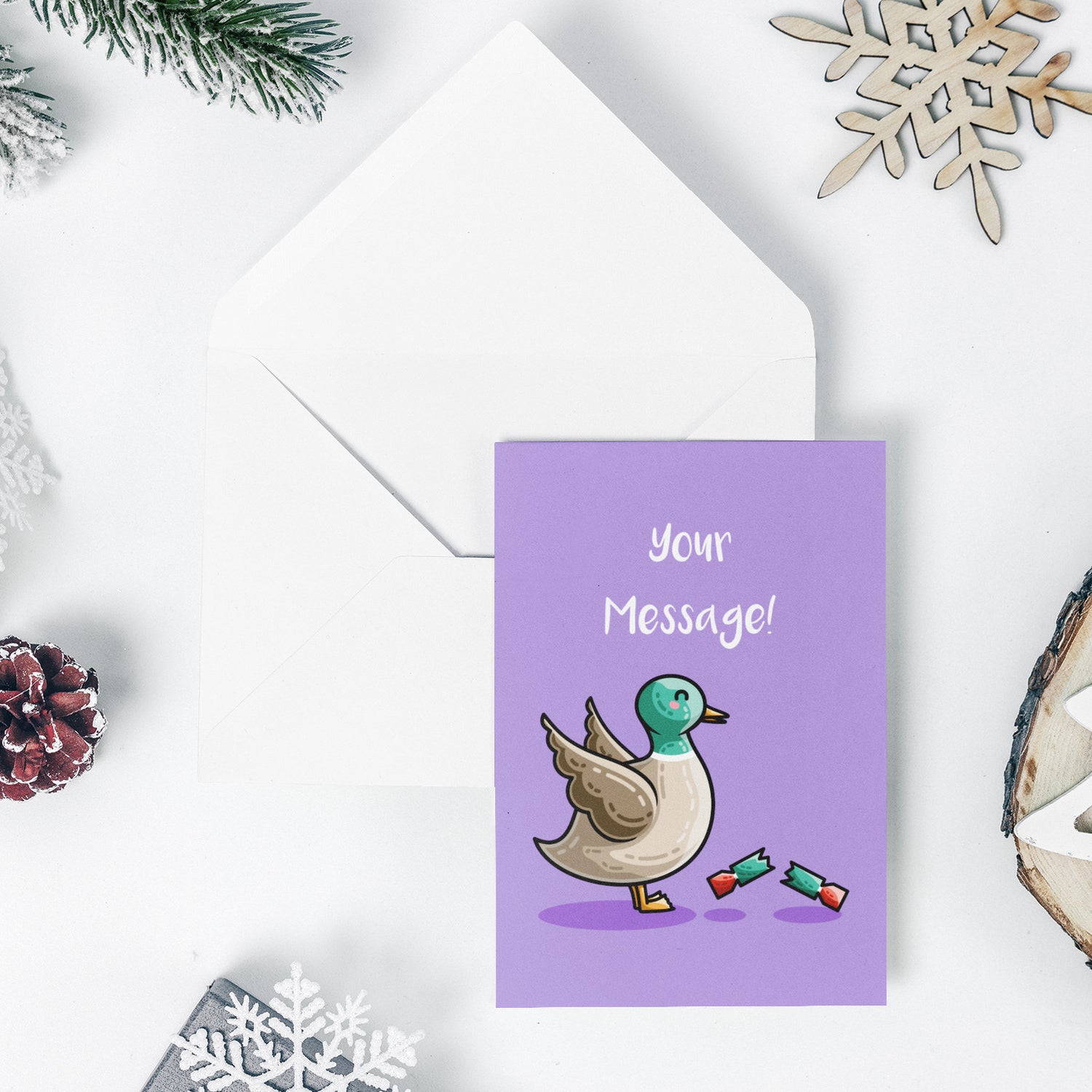 An open white envelope beneath a green greeting card with a design of a cute mallard duck with wings up in alarm at a pulled red and green Christmas cracker at its feet and the words Christmas quacker in white above