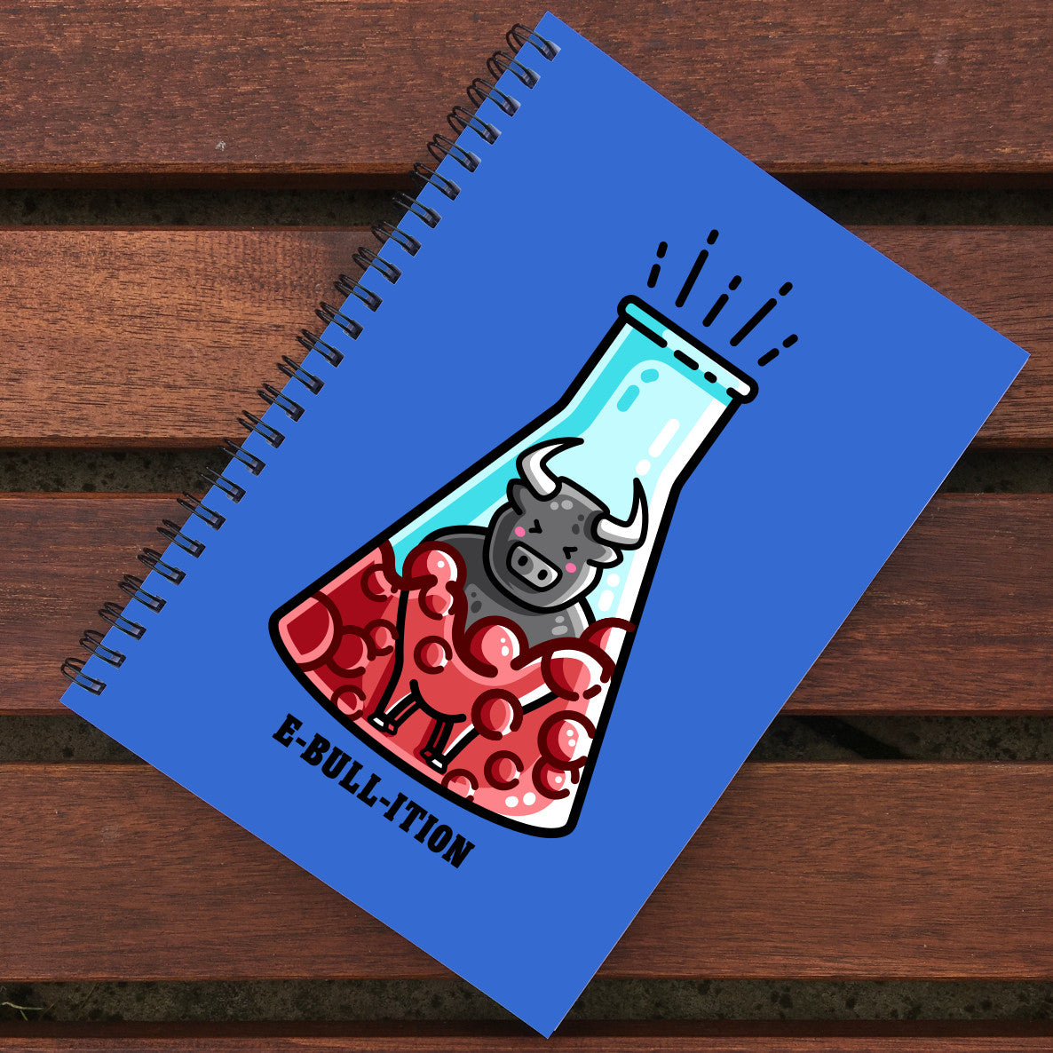 Blue notebook with black spiral binding lying on wooden slats, notebook design is of a kawaii cute bull in a boiling flask of red liquid with 'e-bull-ition' beneath.