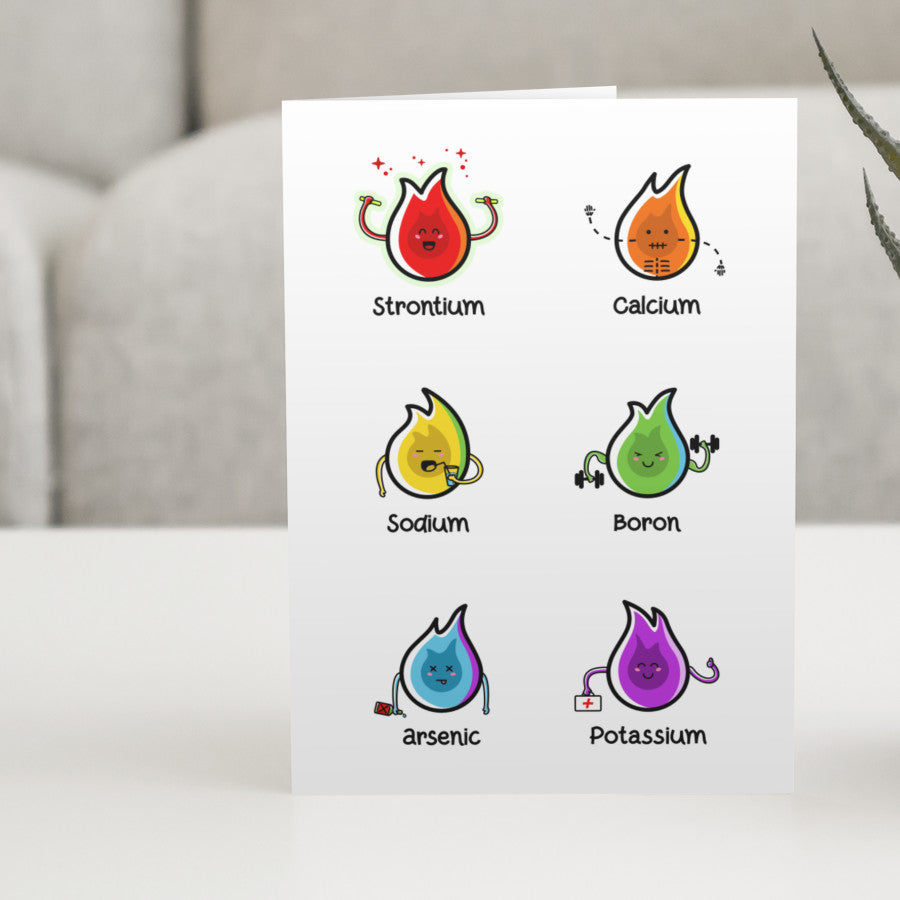 A white greeting card standing on a white table with a design of six cute coloured chemistry element flames.