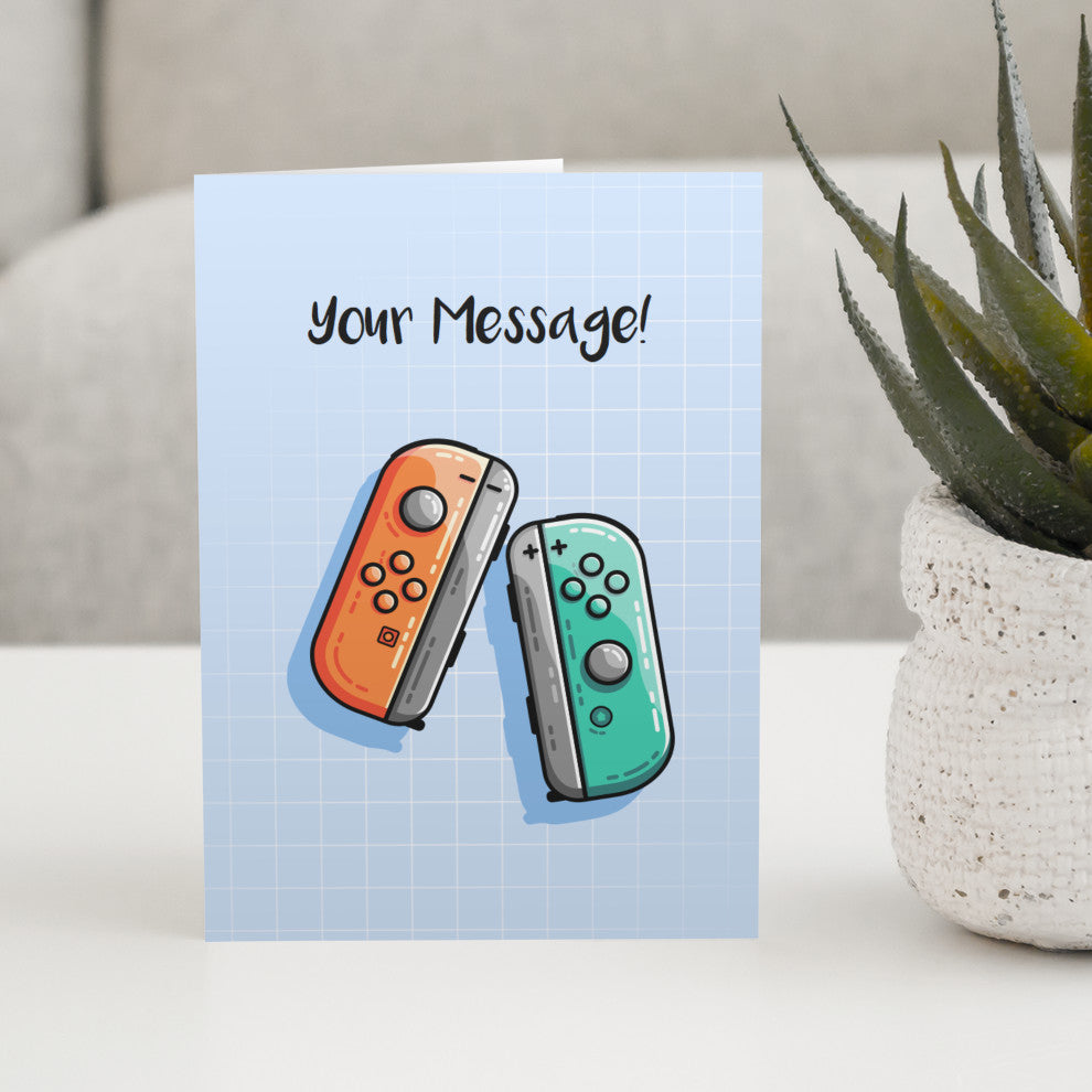 A blue greeting card standing on a white table with a design of an orange and a turquoise game controller and the words your message written above